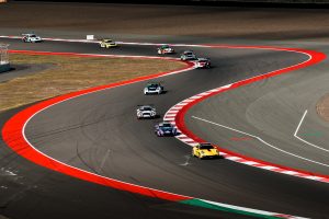 An overhead photograph of a group of eight Porsche 911 GT3 Cup cars driving on a formation lap being lead by a yellow Porsche 911 Carrera S safety car.