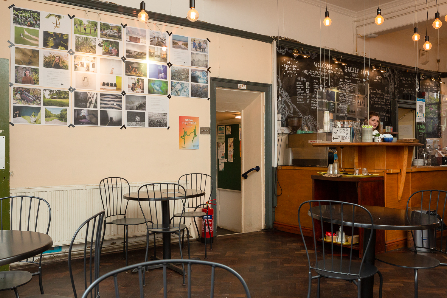 Photograph in the Gallery Café at St. Margaret's House, showing an exhibition of photographs printed on newsprint, tables and chairs in the foreground, and the café counter to the right.