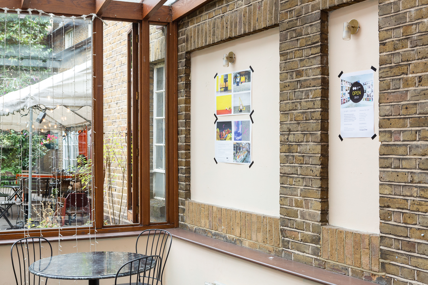 Photograph in the Gallery Café at St. Margaret's House, showing an exhibition of photographs printed on newsprint, tables and chairs in the foreground