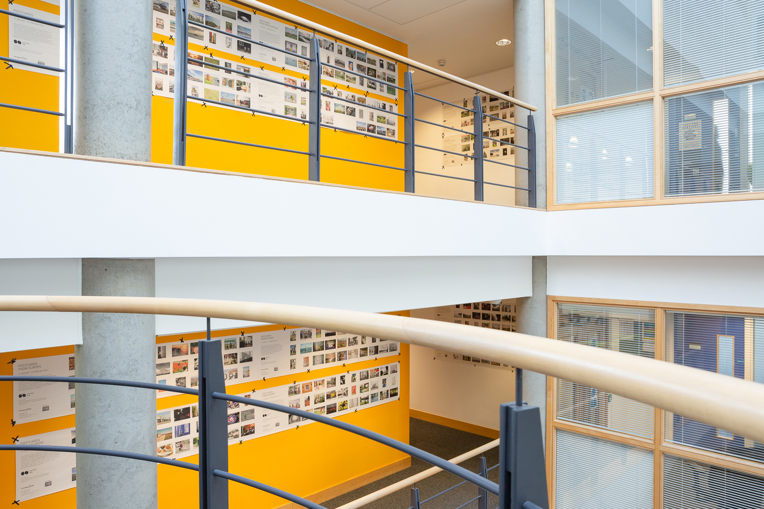 Installation photograph of 'Postcards from Europe' exhibition, showing postcard-sized photographs printed on long strips of newsprint, taped to yellow walls and white walls further in the distance, across two floors with concrete pillars and railings in front and windows to the right.