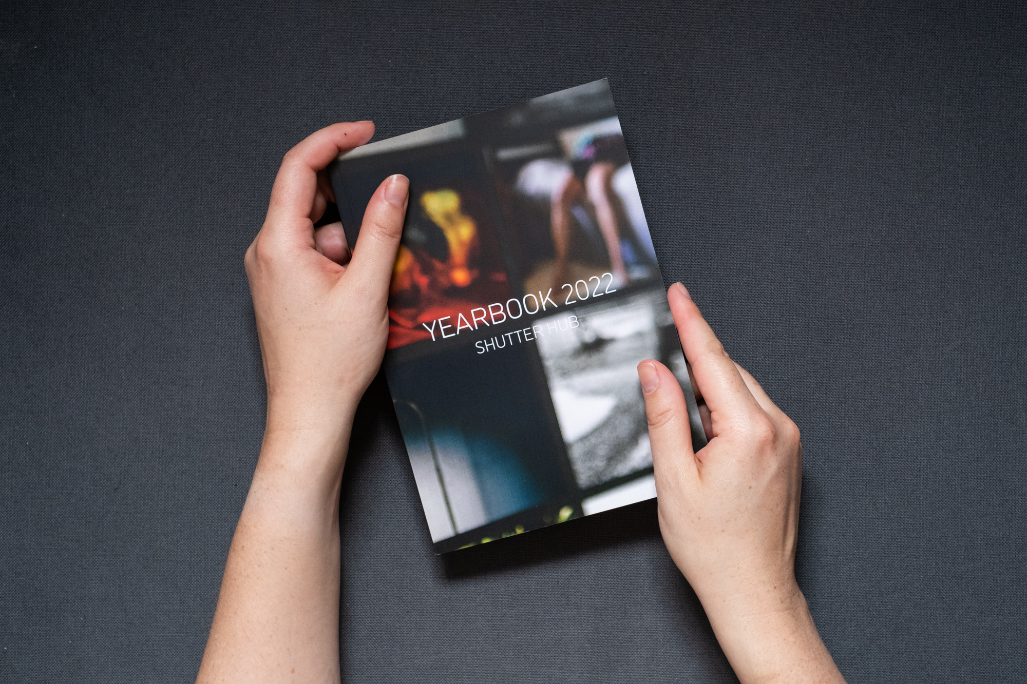 Photograph of hands holding a book against a dark grey fabric background. The front cover is shown, with text reading 'YEARBOOK 2022, SHUTTER HUB' and an out of focus photograph of images from the 2021 YEARBOOK online exhibition on a monitor in the background.